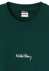 T-shirt en coton vert avec un col rond côtelé. Présente un logo "Keith Haring" brodé en blanc sur le devant. Texture douce, coupe ample.