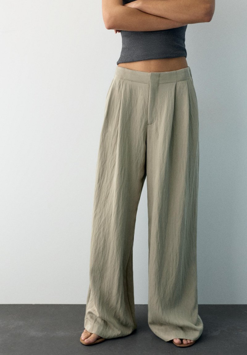 Personne debout, les bras croisés, portant un pantalon beige à jambes larges, un haut court foncé et des sandales, sur un fond uni.