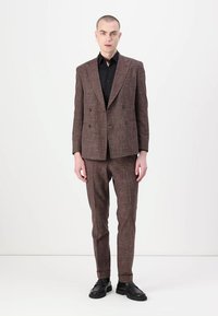 Sacou dublu cu carouri maronii, cu pantaloni slim asortați, purtat peste o cămașă neagră, având linii ascuțite și o croială ajustată.