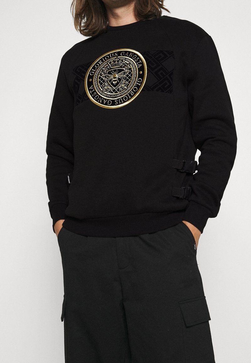 Homme portant un sweat-shirt noir avec un logo circulaire doré représentant une abeille et le texte « Glorious Gangsta », associé à un pantalon noir avec des poches latérales.