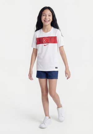 Niña joven sonriendo, vistiendo una camiseta blanca de Nike con la bandera de Turquía, pantalones cortos azul marino y zapatillas blancas, caminando hacia adelante sobre un fondo blanco.