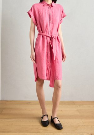 Femme portant une robe rose boutonnée à manches courtes et longueur genou, avec une ceinture nouée à la taille, et des ballerines Mary Jane noires sur un sol en bois.