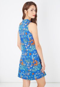 Joven mujer con cabello largo y castaño, vistiendo un vestido sin mangas azul con patrones florales naranjas y verdes, mirando por encima de su hombro.