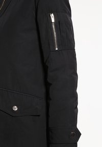 Černá voděodolná bunda má kapsu na zip na rukávu, zapínání na druky a strukturovanou látku s matným povrchem.