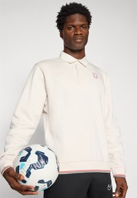 Mand iført en hvid langærmet polo og sorte bukser holder en fodbold foran en ensfarvet baggrund.