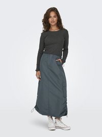 Langer, grauer, hochgeschnittener Rock mit Kordelzugdetails und seitlichen Taschen, kombiniert mit einem dunklen Langarmoberteil und weißen Sneakers.