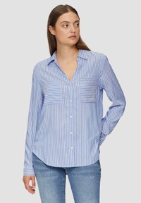 Chemise à rayures bleues et blanches avec un décolleté en V, des manches longues et deux poches avant. Tissu doux avec une texture lisse.