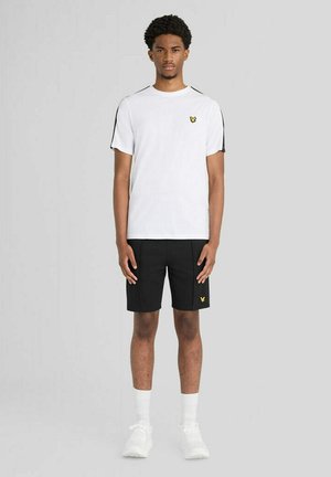 Männliches Model trägt ein weißes T-Shirt mit schwarzen Streifendetails und schwarze Shorts mit passendem gelbem Logo, steht vor einem schlichten grauen Hintergrund.