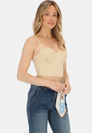 Kobieta o blond włosach, ubrana w beżowy króciutki top bustier i niebieskie dżinsy z wysokim stanem, trzymająca w rękach wzorzysty szal.