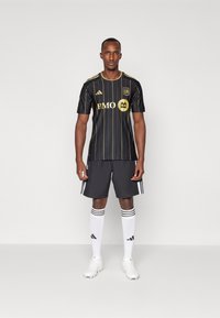 Maglia da calcio nera con strisce dorate e logo BMO, abbinata a pantaloni corti neri, calze bianche con strisce nere e scarpini bianchi.