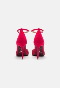 Crvene suede štikle s tankom narukvicom oko gležnja, s ukrasnom zakovicom na peti; dizajn s oštrim vrhom i stiletto oblikom.
