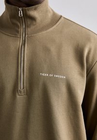 Felpa verde oliva con colletto alto e mezzo zip. Presenta la scritta "TIGER OF SWEDEN" stampata in bianco sul davanti. Tessuto morbido e testurizzato.