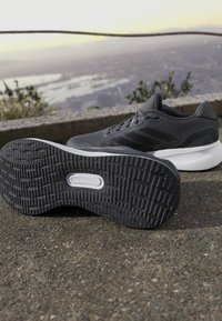 Scarpa da ginnastica grigia con suola nera testurizzata, caratterizzata da una parte superiore liscia con accenti sottili e una intersuola bianca, posata su una superficie pavimentata.