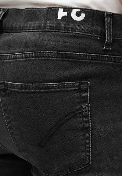 Fekete farmernadrág hátzsebének közeli képe, díszítő varrással és fém "D" logó részlettel a derékrész közelében.