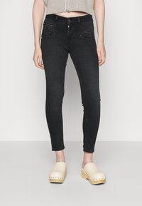 Jeans skinny negros de denim, con bolsillos frontales con cremallera y acentos metálicos. Combinados con un top corto y zuecos de madera clara.