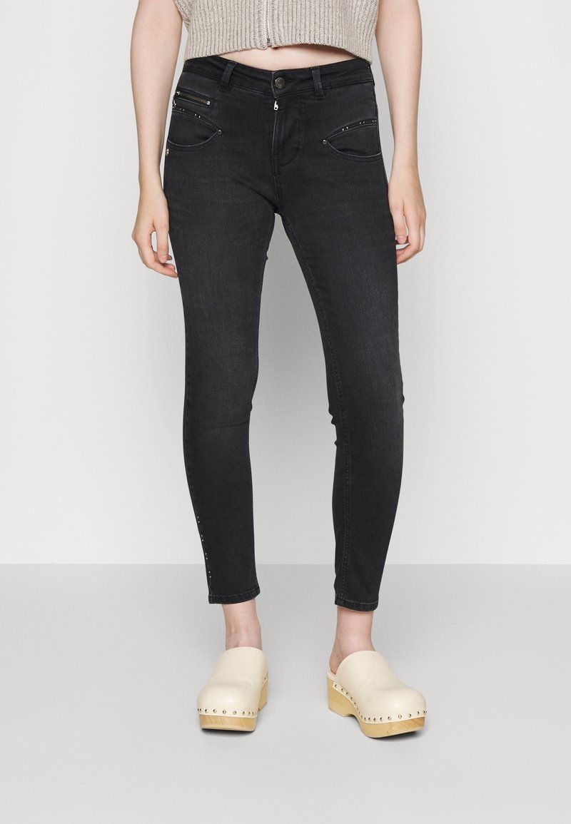 Jeans skinny negros de denim, con bolsillos frontales con cremallera y acentos metálicos. Combinados con un top corto y zuecos de madera clara.
