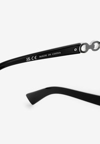 Montura de gafas de sol negras con textura suave. Cuenta con herrajes plateados en la bisagra. Marcadas con etiquetas "CE" y "HECHO EN CHINA".