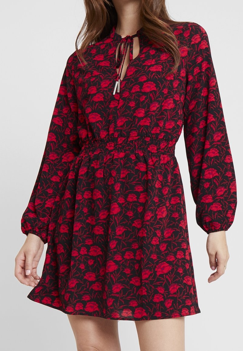 Femme portant une robe noire à manches longues avec un motif floral rouge, taille froncée et col à nouer avec des embouts métalliques.