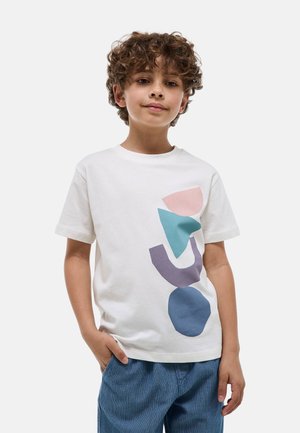 Garçon aux cheveux bouclés portant un T-shirt blanc avec des formes abstraites bleu, violet et rose, associé à un pantalon bleu, mains dans les poches.
