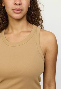 Beež ribakujuline pitsidop, millel on ümar kaelus ja laiad rihmad, omades sujuvat tekstuuri ja keha lähedast disaini.