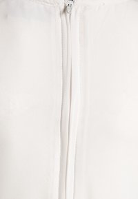 Tela blanca, de textura suave, con un cierre central de cremallera. La cremallera es metálica, con un acabado limpio en los bordes de la costura.