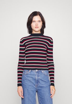 Ribbestrikket genser med horisontale striper i svart, rødt og grønt. Har høy hals og tettsittende lange ermer, kombinert med blå jeans.