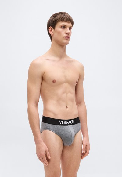 Jeune homme aux cheveux bruns courts portant un slip gris avec une ceinture noire portant la marque "Versace", debout sur un fond clair uni.