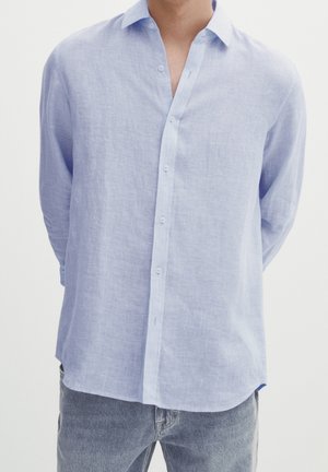 Uomo che indossa una camicia di lino azzurro chiaro a maniche lunghe con bottoni e colletto, abbinata a jeans denim grigio chiaro, in piedi con le mani dietro la schiena.