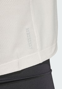 Camiseta atlética blanca, texturizada y con un diseño de malla, que presenta un dobladillo cosido y el logo "AEROREADY" en gris claro. Combinada con pantalones de tela negros.