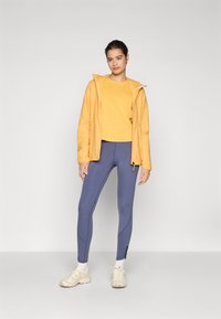 Veste zippée jaune par-dessus une chemise jaune, associée à des leggings bleus et des chaussures de sport beiges. Tissu lisse et coupe décontractée.