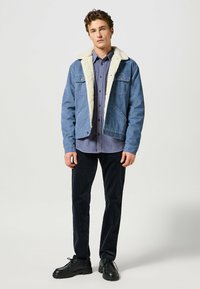 Denimjacka med faux fur-krage, ljusblå färg, med två bröstfickor och knapplås framtill, bärs över en grå skjorta och svarta kordbyxor.