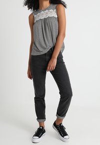 Wzorzysty bezrękawnik z koronkowymi detalami, zestawiony z ciemnoszarymi dżinsami podwiniętymi na nogawkach i czarnymi sneakersami z białymi podeszwami.