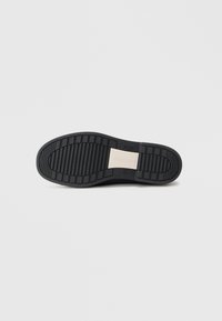 Semelle de chaussure en caoutchouc noir avec des rainures horizontales et verticales pour une meilleure traction, dotée d'un accent en métal au centre.