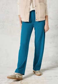 Tealblauwe broek van rekbaar materiaal met een rechte snit. Gecombineerd met beige sportschoenen en een crèmekleurig fleece vest.
