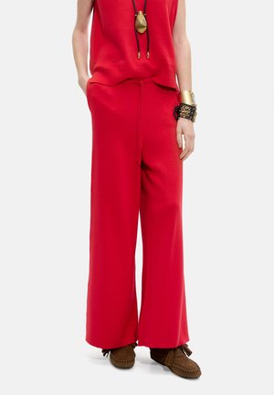 Kobieta w czerwonych spodniach o szerokich nogawkach i bezrękawniku, nosząca brązowe mokasyny oraz złoto-czarne bransoletki, stojąca z rękami w kieszeniach.