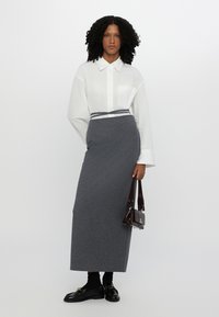 Larga falda gris de tejido suave, combinada con una camisa blanca de botones. Presenta una cintura definida con un acento de rayas oscuras. Bolso marrón y mocasines.