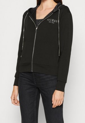 Vrouw draagt een zwarte hoodie met ritssluiting en de tekst "GUESS EST. 1981" op de borst, gecombineerd met een donkere spijkerbroek, staand tegen een effen achtergrond.