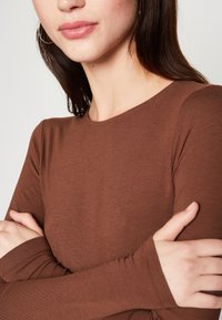 Smeđi dugi rukav s okruglim izrezom. Ima glatku, rastezljivu tkaninu i prilagođene rukave. Model drži ruke prekrižene, pokazujući manžete.