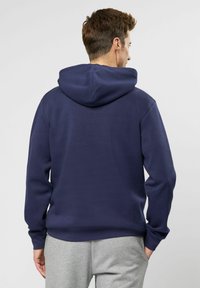 Sudadera azul marino con capucha ajustable, fabricada con un material suave, que presenta un corte relajado y puños acanalados. Pantalones de chándal grises parcialmente visibles.