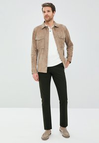 Beige suède jack met twee voorzakken, witte blouse met lange mouwen, slim-fit donkergroene broek en lichtgrijze instapschoenen.