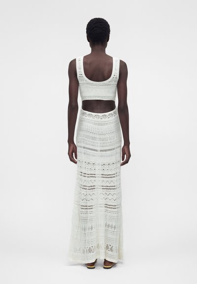 Γυναίκα με κοντά μαλλιά που φοράει λευκό, αμάνικο, πλεκτό κορμάκι και μακριά πλεκτή φούστα που ταιριάζει, στέκεται με την πλάτη προς τον θεατή.
