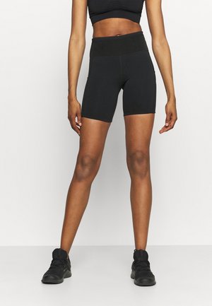 Femme portant un short cycliste noir taille haute, un soutien-gorge de sport noir et des chaussures de sport noires, debout devant un fond clair uni.