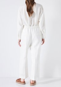 Vit linnebyxor med avslappnad passform, elastisk midja och sidofickor. Bärs med bruna sandaler med remmar. Endast bakifrån.