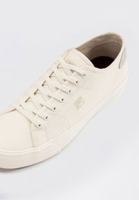 Sneaker in tela crema con lacci bianchi, suola in gomma e una parte superiore testurizzata. Presenta un logo discreto sul lato e un accento sul tallone in contrasto.