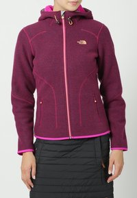 Kobieta w fioletowej bluzie z zamkiem błyskawicznym, z różowymi akcentami, oraz czarną pikowaną spódnicą, stojąca na jednolitym tle.