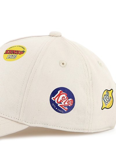 Casquette de baseball beige ornée de plusieurs écussons circulaires en bleu, jaune et rouge, affichant les logos "Kenzo" et "1970". Tissu texturé.