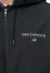 Κοντινό πλάνο ενός μαύρου αθλητικού μπουφάν New Balance με φερμουάρ, κουκούλα με κορδόνια και εμφανές λογότυπο στο στήθος.