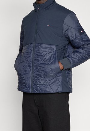 Uomo che indossa una giacca trapuntata blu navy con cerniera frontale e tasche, in piedi con le mani nelle tasche davanti a uno sfondo semplice.