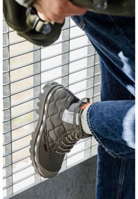 Graue gepolsterte Knöchelstiefel aus strukturiertem Stoff, Gummisohle, reflektierende Akzente und orangenen Tag. Getragen mit blauen Jeans und Wollsocken.