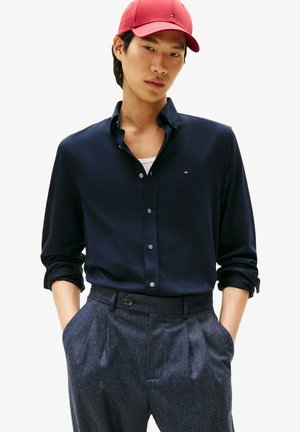 Young man wearing a red cap, navy button-up shirt with logo, gray pleated trousers, hands in pockets, looking slightly downward with neutral expression.